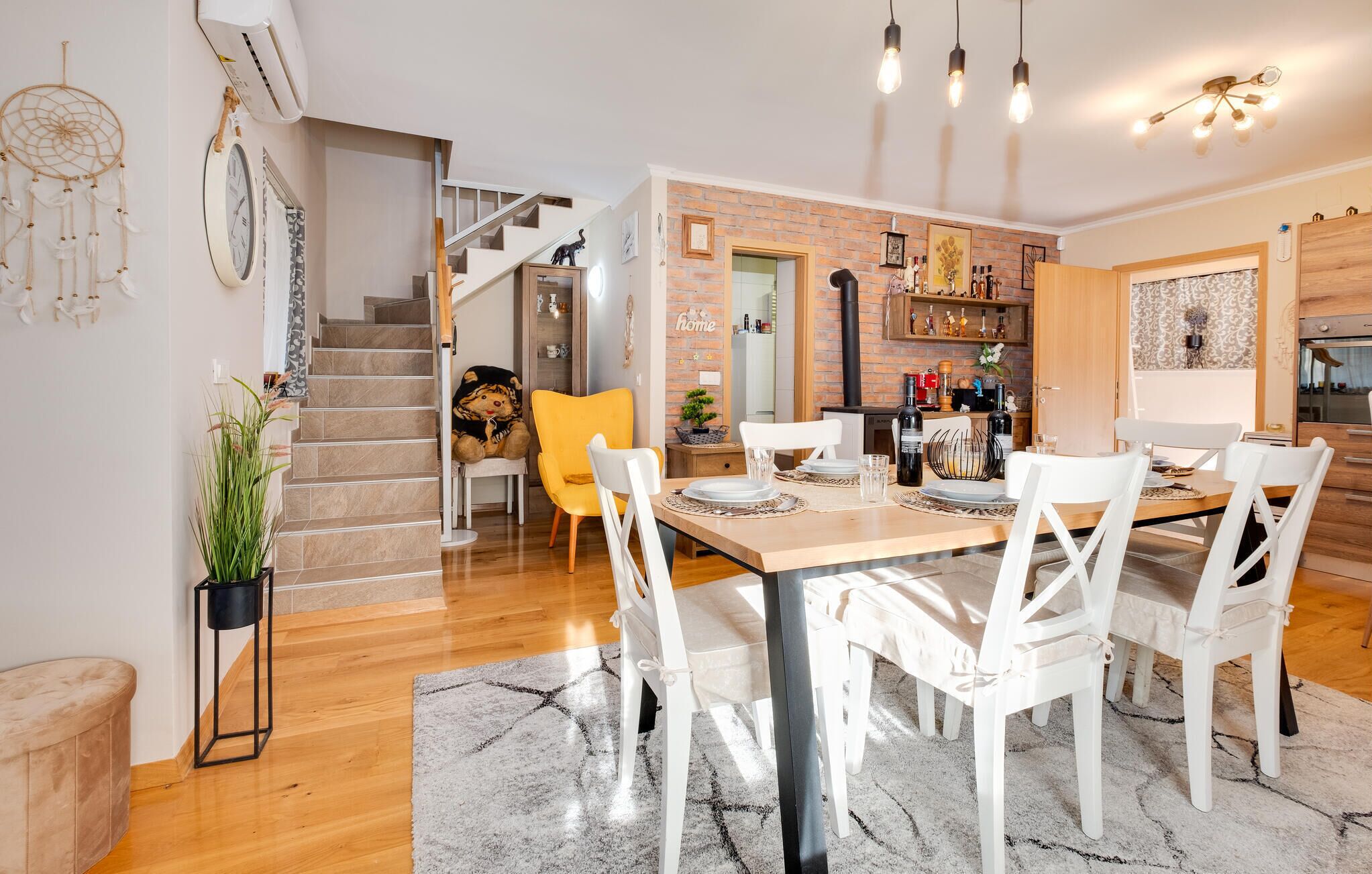 Cozinha moderna com parede de tijolos, armários de madeira e área de jantar com cadeiras brancas e mesa de madeira.
