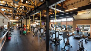 Ein Fitnessraum mit unterschiedlichen Trainingsgeräten, Holzbalken an der Decke und großen Fenstern.