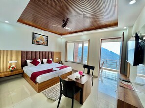 Ein modernes Hotelzimmer mit einem Holzkopfteil, einem großen Bett mit rot-weißer Bettwäsche, einem Schreibtisch mit Stuhl und Blick auf Berge durch die Fenster.