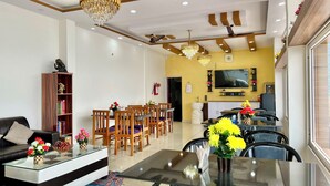A modern living room with a glass coffee table, a sofa, and a dining area with chairs and a table. There is a television mounted on the wall, a bookshelf, and a chandelier. - Dhurva Hotel and Resort (Lansdowne)