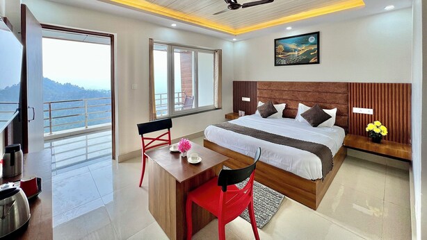 A modern hotel room with a large bed, a desk, and a balcony with a view of the mountains. - Dhurva Hotel and Resort (Lansdowne)