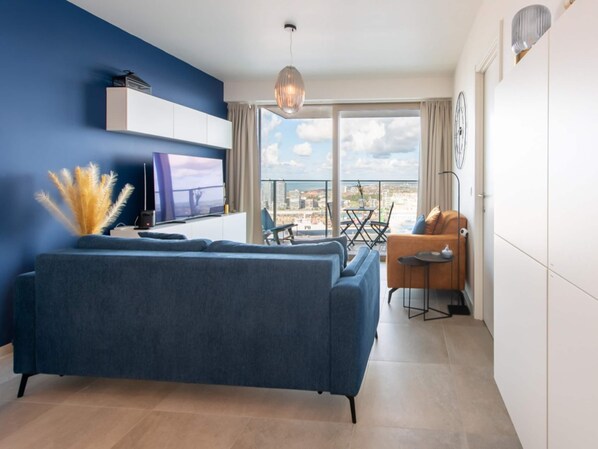 A modern living room with a blue sofa, a flat-screen TV, and a balcony with a city view. - Central Apartment in Ostend Near Beach (Ostend)