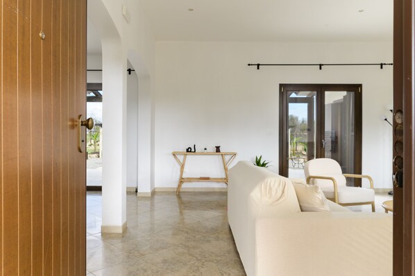 Ein modernes Wohnzimmer mit einer Couch, einem Couchtisch und einem Sessel. Eine Holztür ist links und eine Glastür führt nach draußen.