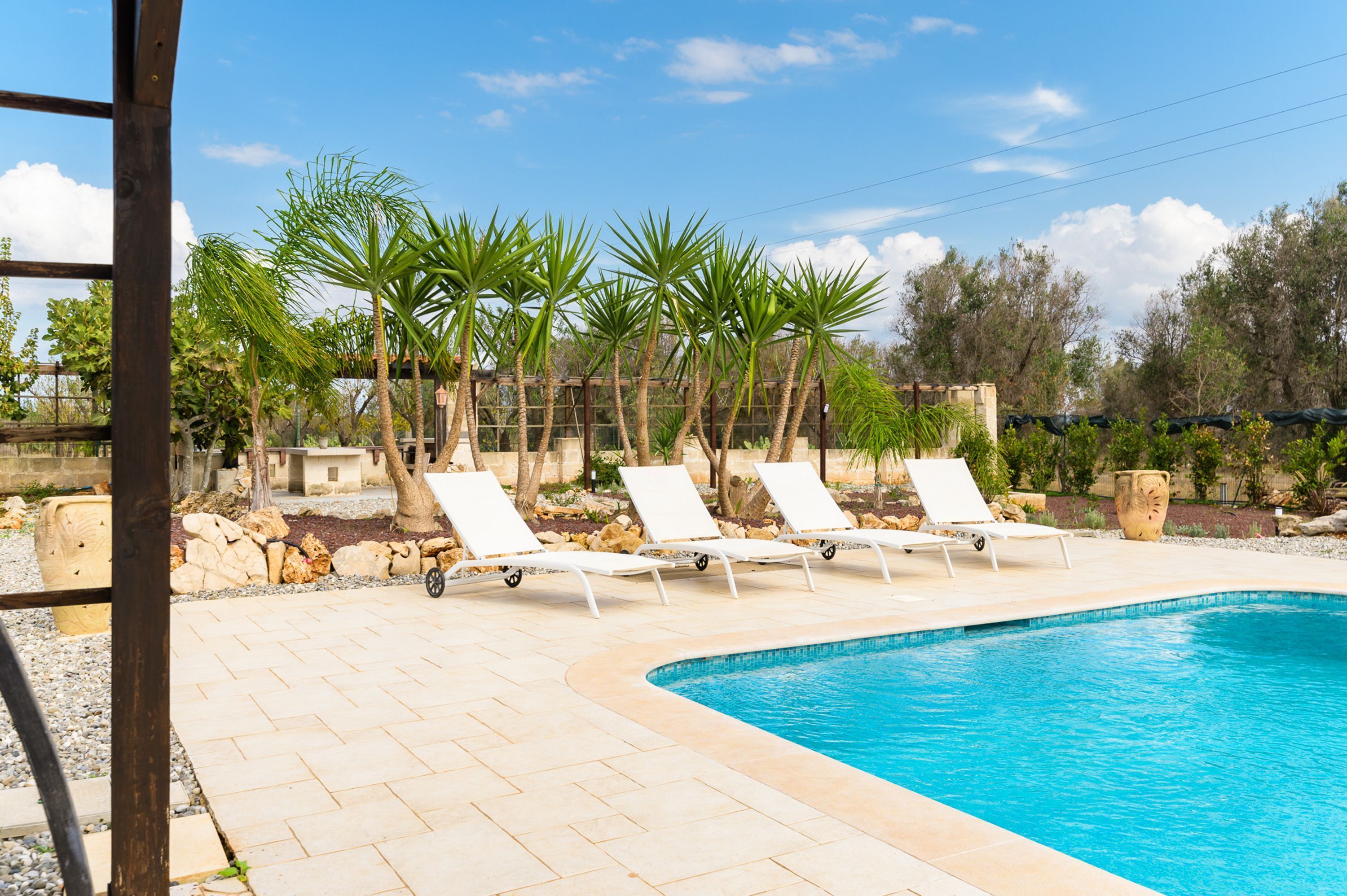 Ein Poolbereich mit weißen Liegestühlen, Palmen und wolkenlosem, blauem Himmel.