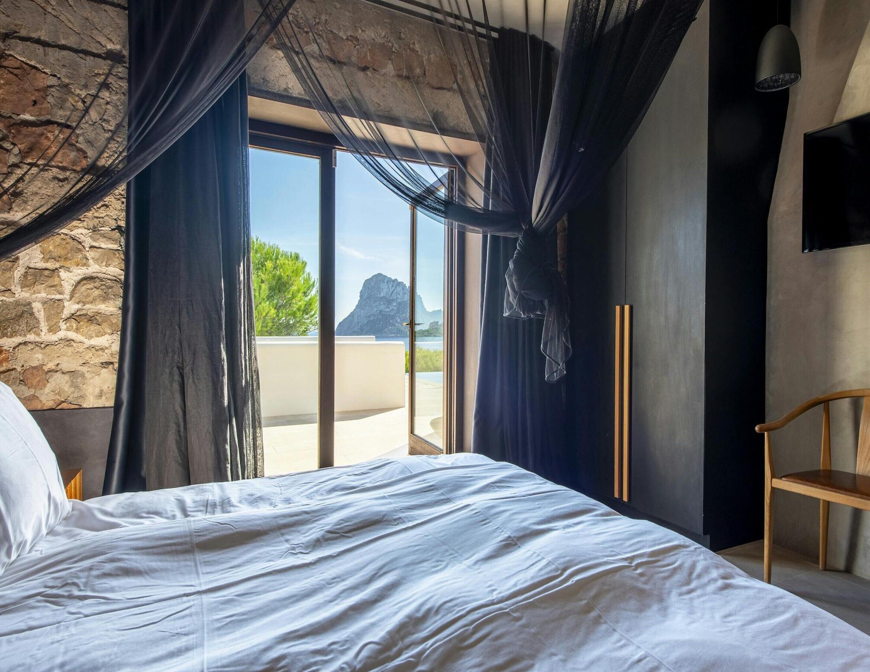 Un dormitorio con una cama grande, una silla de madera, un televisor montado en la pared y vistas a la montaña a través de un ventanal con cortinas.
