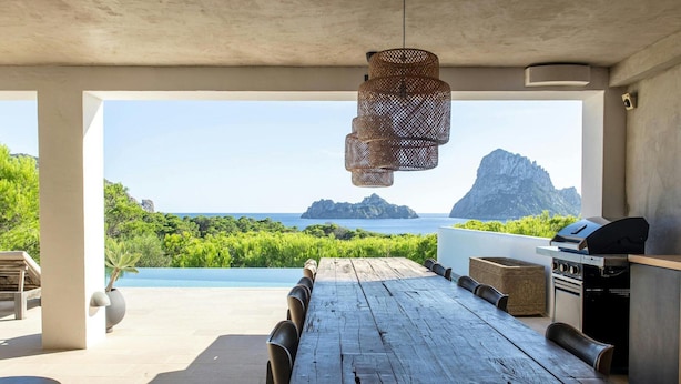 An outdoor dining area with a rustic wooden table and chairs, a hanging light fixture, and a view of the ocean and mountains.