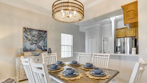Un coin repas avec une table en bois dressée pour six personnes, des chaises blanches, un lustre et un tableau accroché au mur.