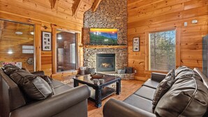 A cozy log cabin interior with a stone fireplace, leather sofas, and a television mounted above it. - Dog Friendly! Private Indoor Pool, Hot Tub, GameRoom, Firepit & Free Passes. (Sevierville)