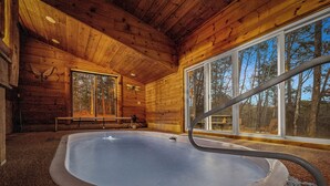 A wooden cabin interior with a hot tub, large windows, and a view of the forest. - Dog Friendly! Private Indoor Pool, Hot Tub, GameRoom, Firepit & Free Passes. (Sevierville)