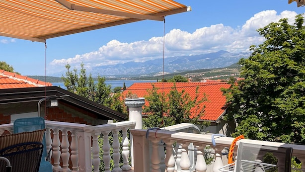 Ein Balkon mit Blick auf einen See und Berge, ein rotes Ziegeldach und ein gestreifter Sonnenschirm.