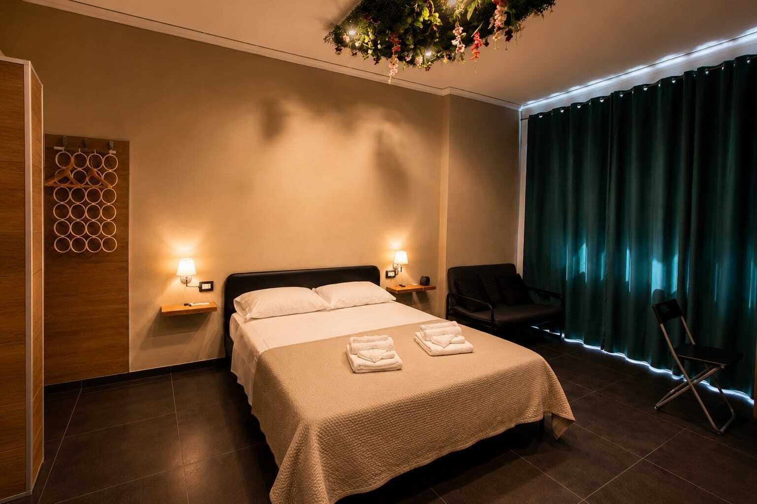 A modern hotel room with a bed, two bedside tables with lamps, a chair, and a large window with curtains.