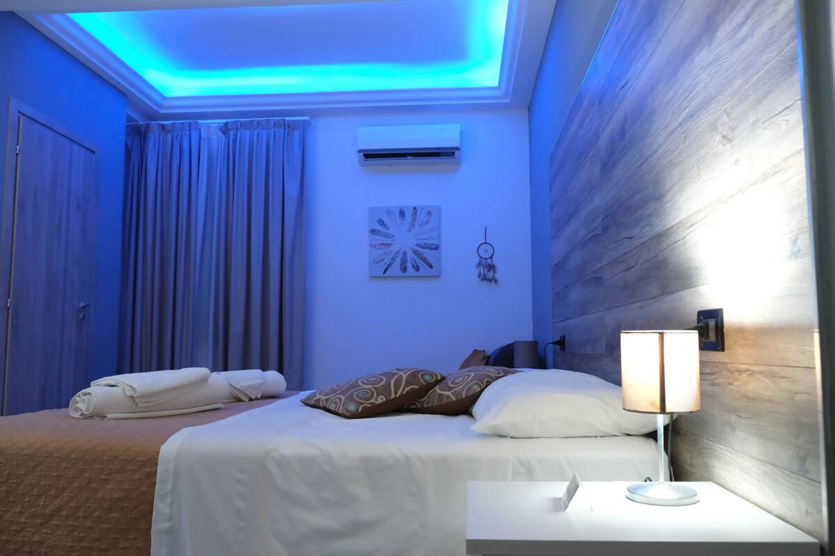 Une chambre d’hôtel avec un lit, une table de chevet avec une lampe, un climatiseur fixé au mur et un plafond doté d’un éclairage bleu.
