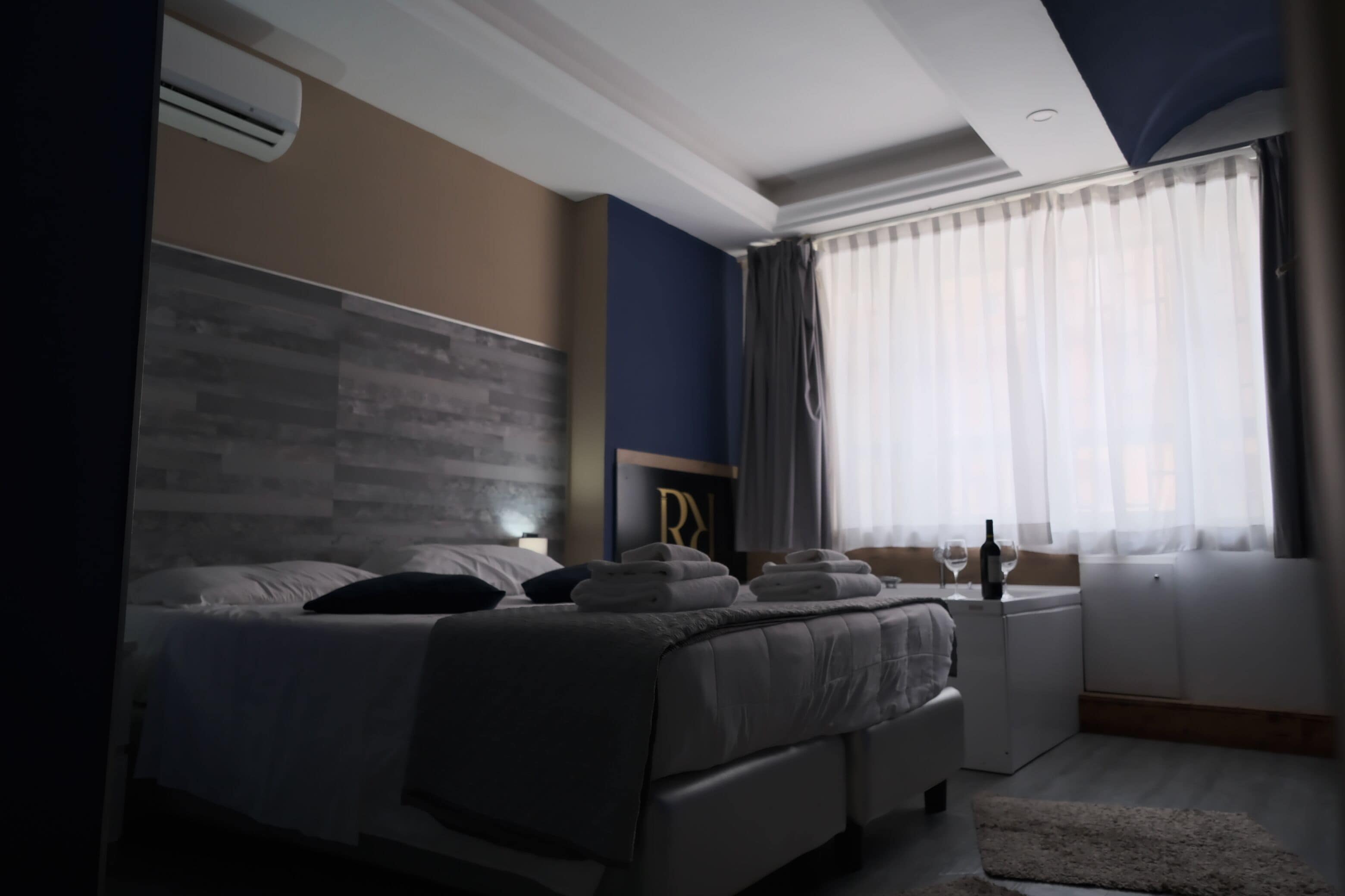 A modern hotel room with a large bed, a bedside table with a bottle and glass, and a window with sheer curtains.