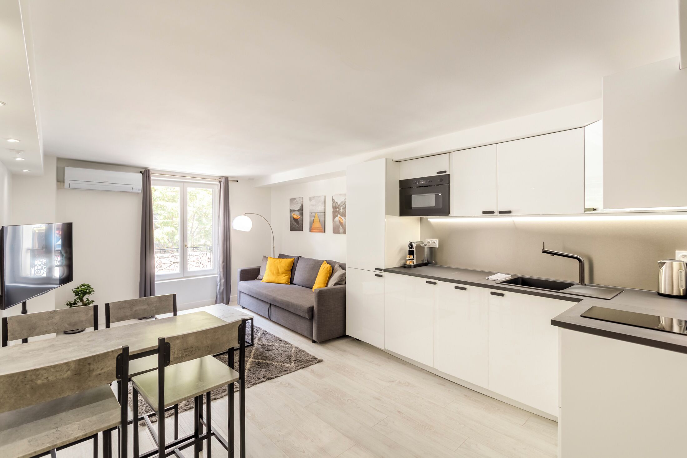 A modern kitchen with white cabinets, a built-in oven, and a sink. There is a dining area with a table and chairs, and a sofa with cushions.