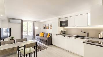 A modern kitchen with white cabinets, a built-in oven, and a sink. There is a dining area with a table and chairs, and a sofa with cushions.