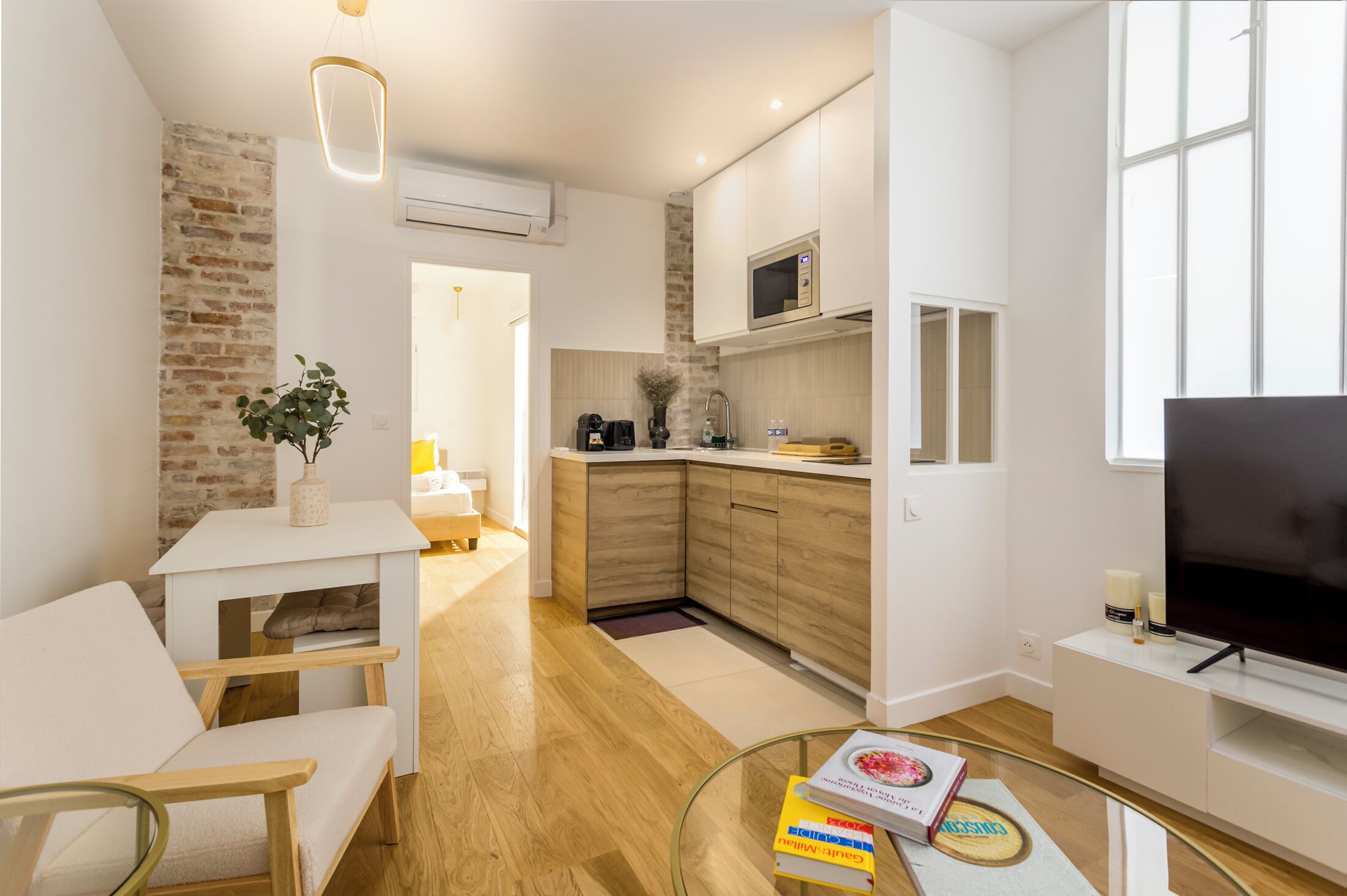 A modern kitchen with white cabinets, a microwave, and a coffee machine. There is a dining area with a white table and chairs, and a TV on a stand.
