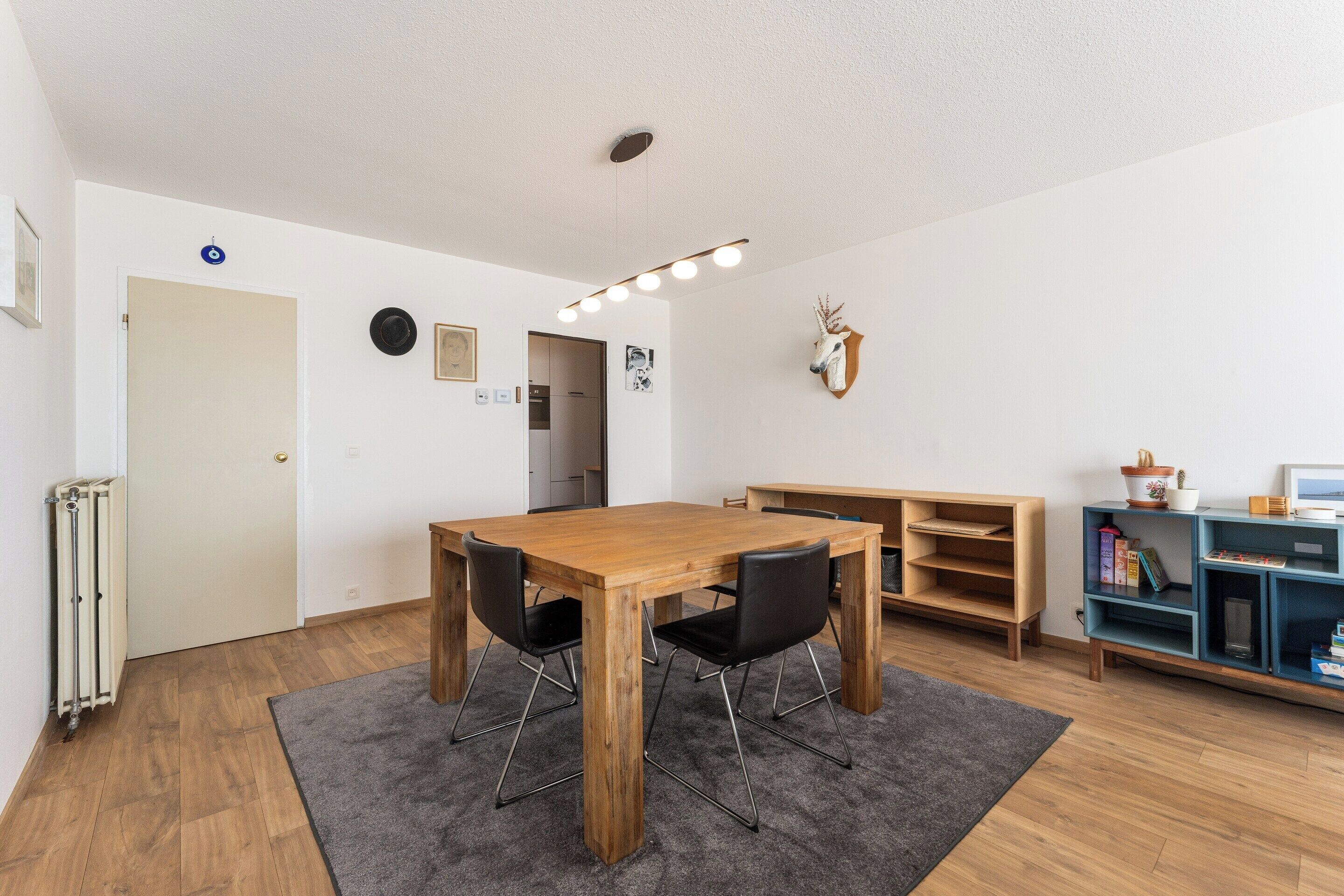 A room with a wooden dining table, two chairs, a grey rug, a radiator, a white door, a wall-mounted light fixture, a shelf with books, and a mounted animal head.