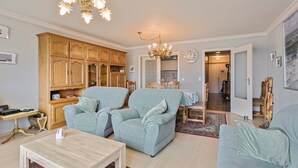 Un salon avec deux fauteuils bleu clair, un meuble en bois, un lustre et un coin repas comprenant une table et des chaises.