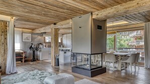 A modern living room with a glass-top fireplace, wooden beams, a dining area, and a view of the city. - LUXURY CHALET CLOSE TO THE VILLAGE (Megève)