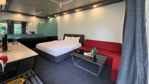 A modern hotel room with a bed, a red sofa, a small table, a desk, and a ceiling fan. - Natures Cove Resort  (Alibag)