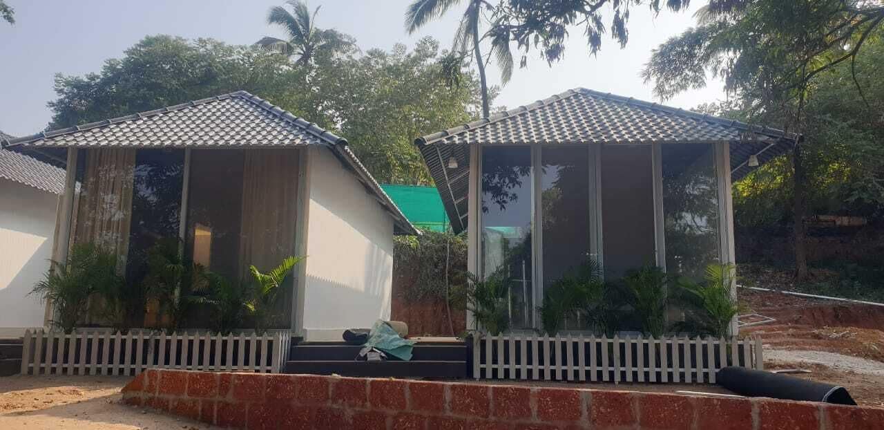 Two small, single-story structures with tiled roofs, surrounded by greenery and a white picket fence.