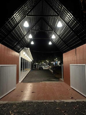 A covered outdoor area with a metal roof, a pathway, and a car parked in the background. - Natures Cove Resort  (Alibag)