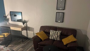 A living room with a brown leather sofa, two yellow cushions, a small black table, a wooden desk with a computer, and a potted plant. - Pine Studio Apartment- Your Own Quiet Space! (Blackpool)
