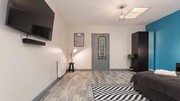 A modern bedroom with a large bed, a flat-screen TV mounted on the wall, a door with a window, and a small side table with a lamp. - Pine Studio Apartment- Your Own Quiet Space! (Blackpool)