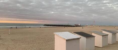 A beach with a row of white beach huts and a pier in the background.