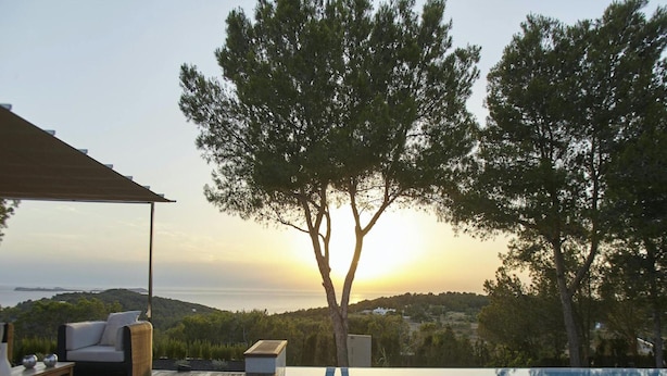 Ein Pool ohne erkennaren Rand mit Holzterrasse, Gartenmöbeln und Blick auf den Sonnenuntergang über dem Meer.