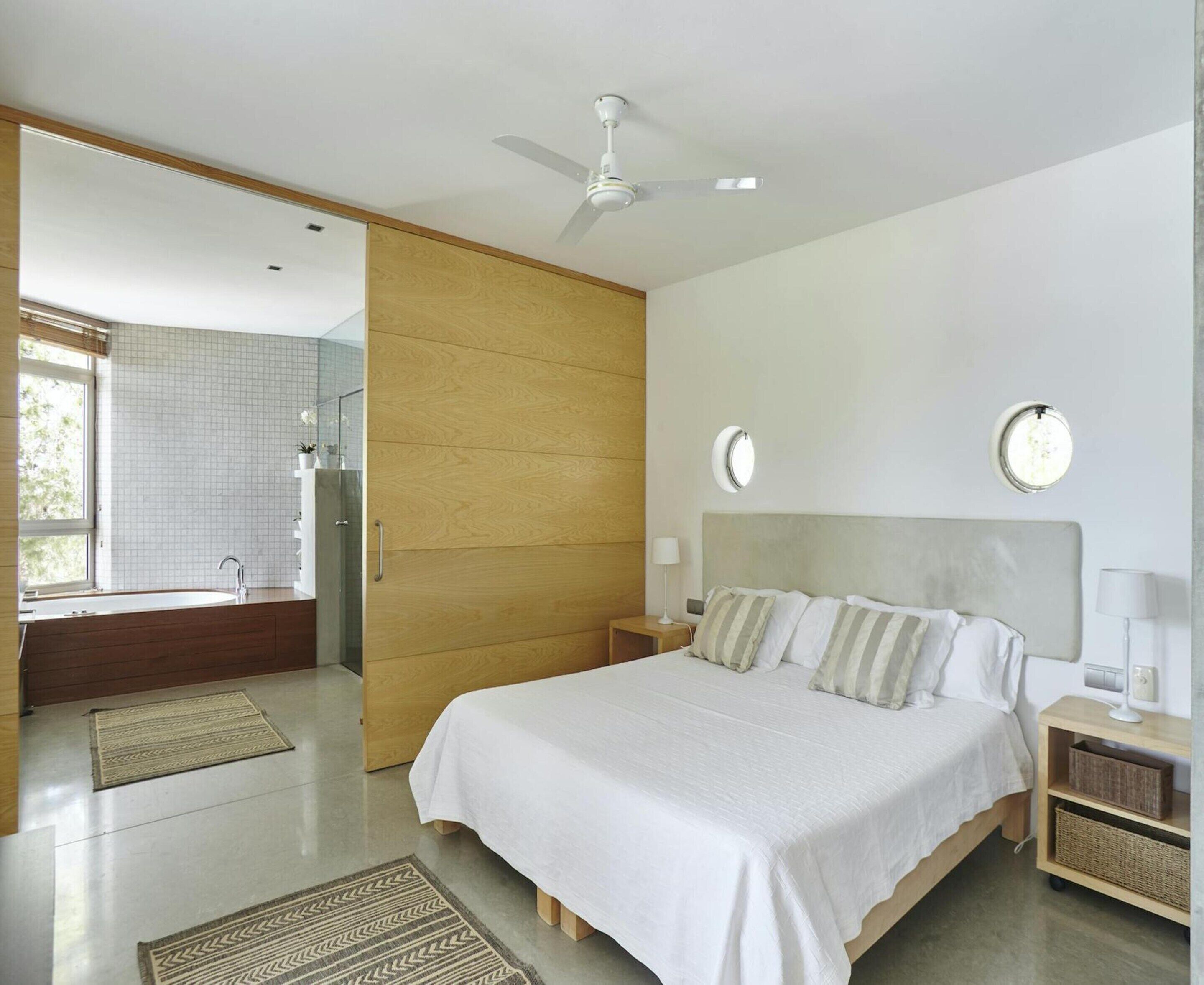 A modern bedroom with a large bed, a bedside table, a nightstand, a ceiling fan, and a window with a view of greenery.