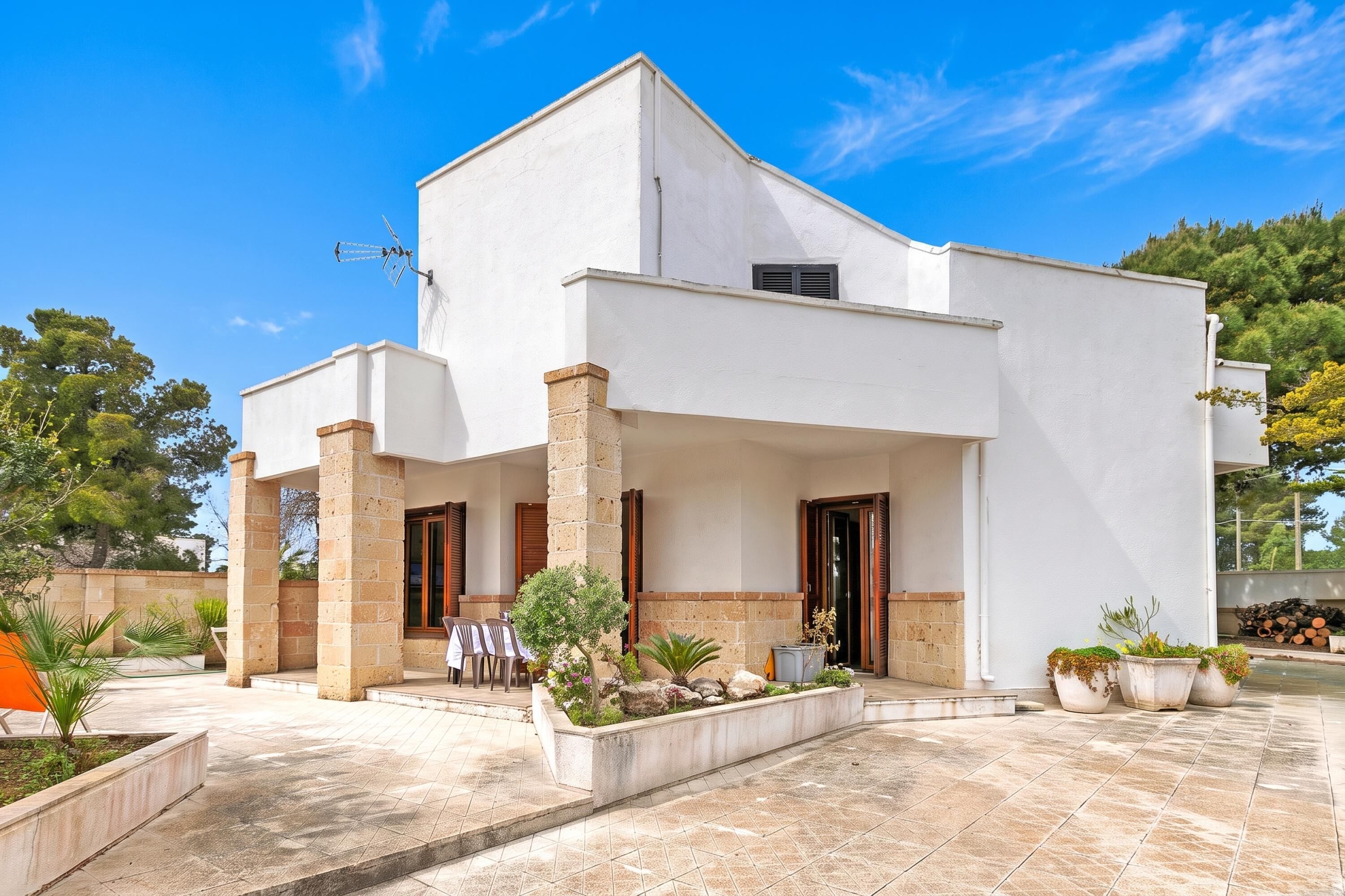 A modern two-story house with a flat roof, a covered patio area with outdoor seating, and a well-maintained garden.