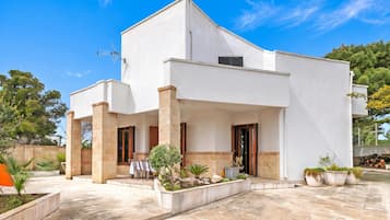 A modern two-story house with a flat roof, a covered patio area with outdoor seating, and a well-maintained garden.