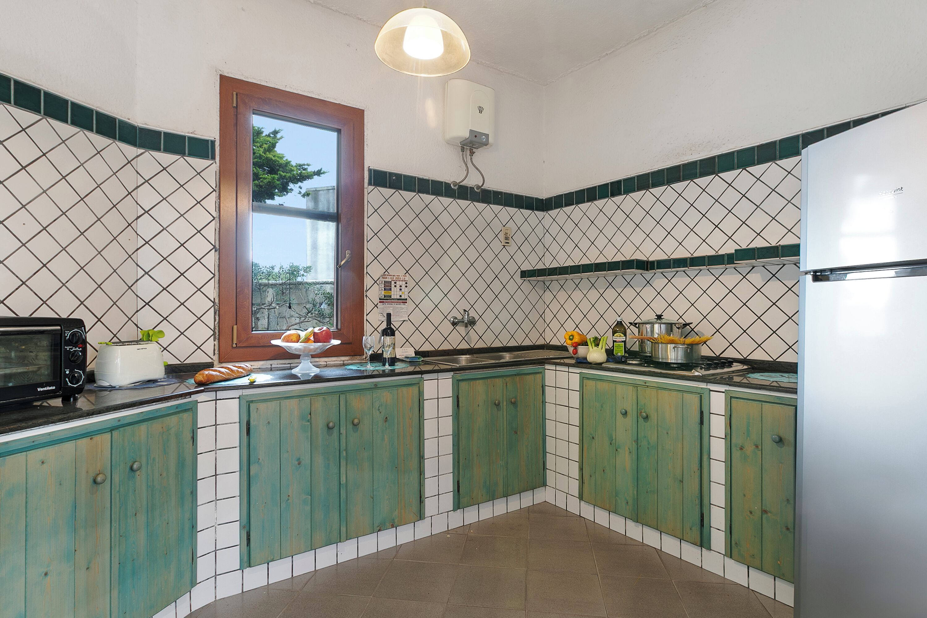 Uma cozinha com armários verdes, micro-ondas, torradeira, pia e janela com vista para árvores.