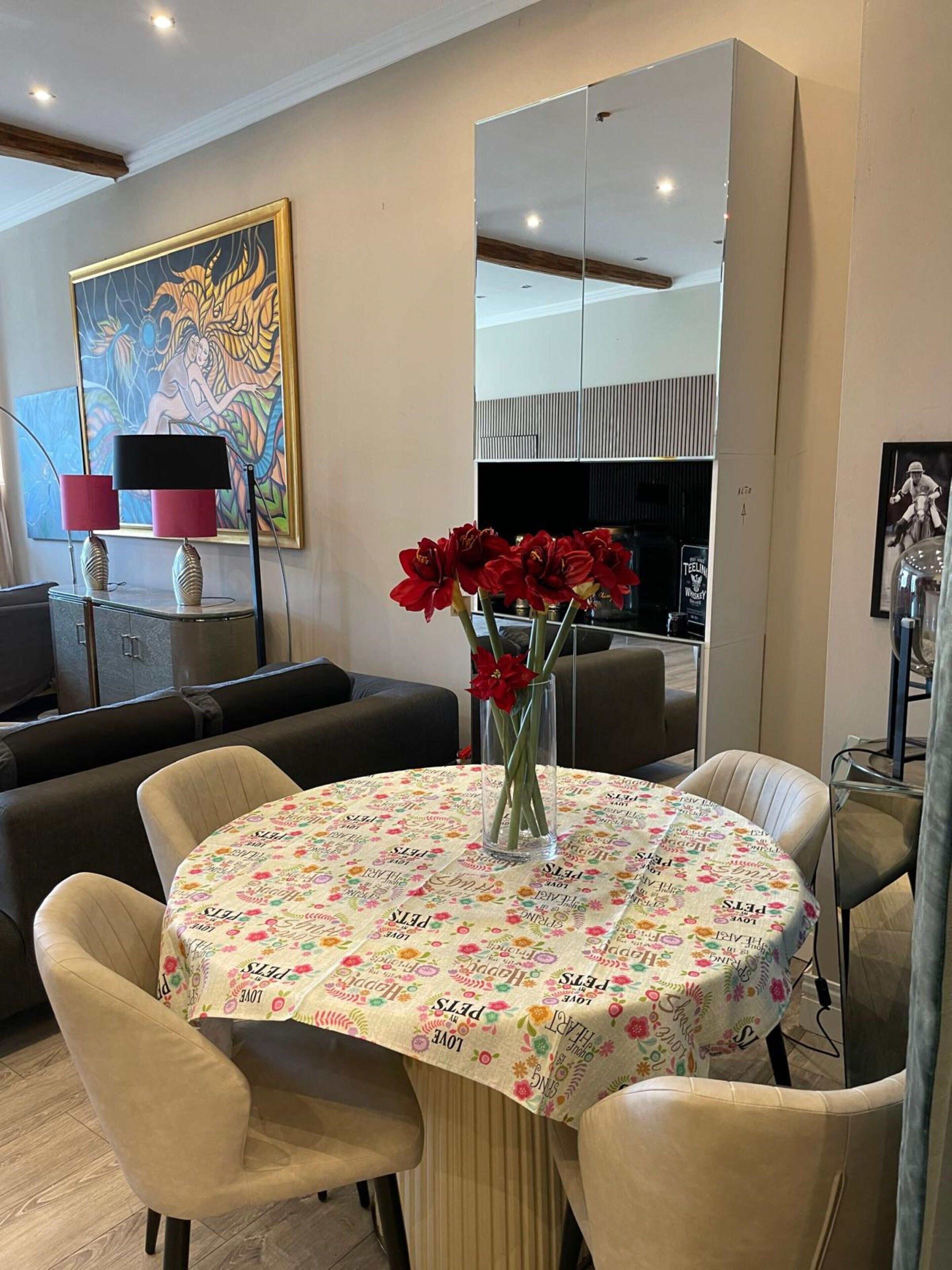 A dining area with a round table covered in a floral tablecloth, surrounded by beige chairs. A vase with red flowers is placed on the table.