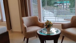 Una habitación con dos sillones beiges, una mesa de centro de vidrio con un florero y vistas a una obra en construcción a través de un ventanal grande.