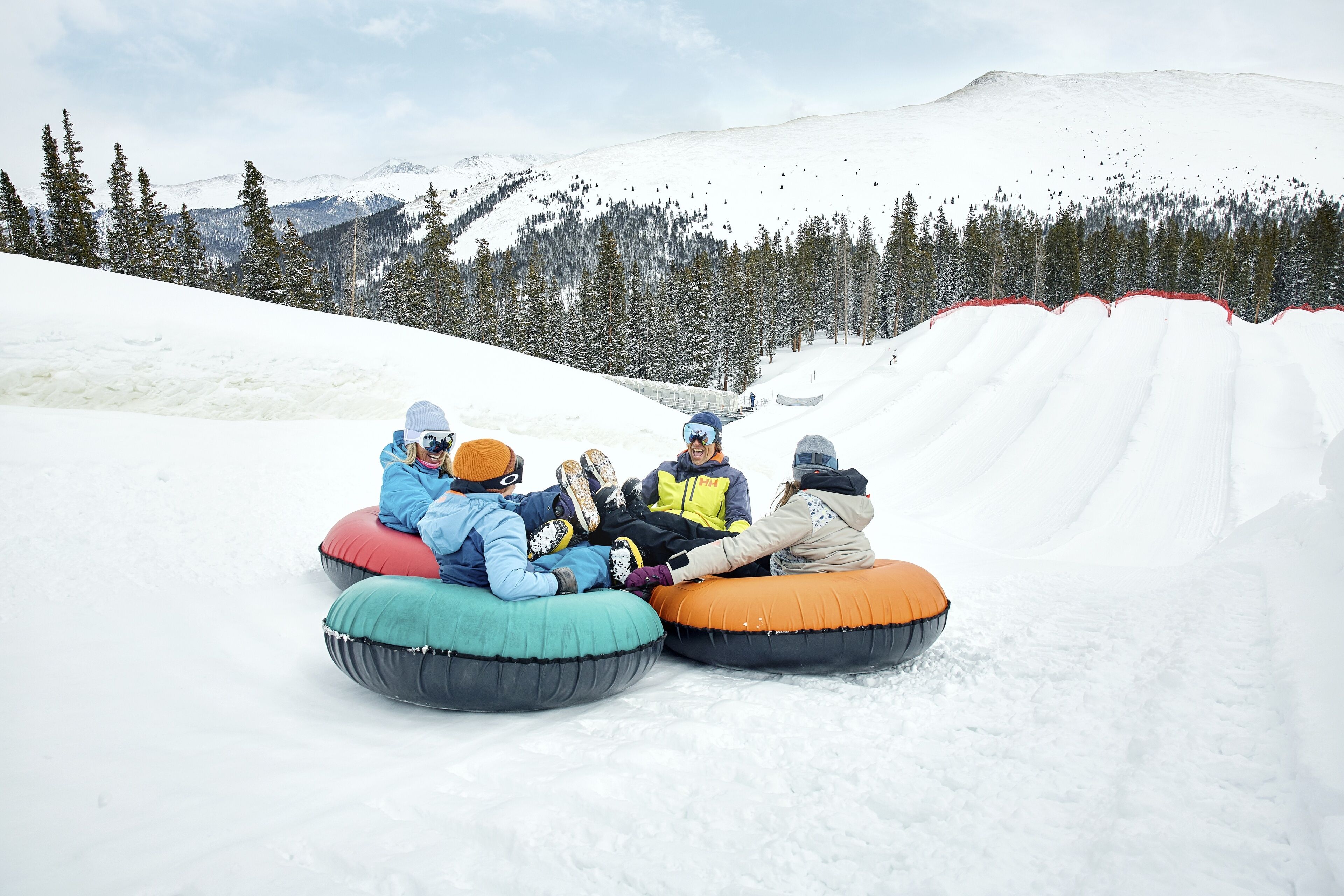 Four people sitting on inflatable snow tubes on a snowy slope with a forested mountain in the background.