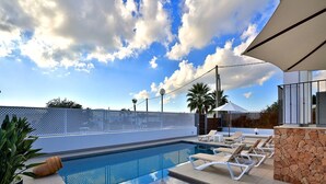 An outdoor swimming pool with lounge chairs and a stone wall, surrounded by a white fence and palm trees under a partly cloudy sky. - Amazing Villa Near Ibiza Town (Santa Eulalia del Rio)