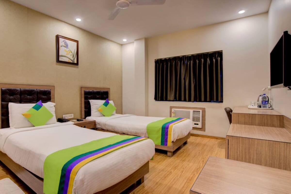 A hotel room with two beds, a desk, a chair, a ceiling fan, a window with curtains, and a wall-mounted TV.