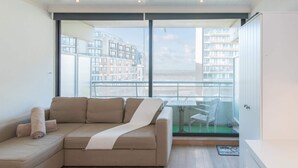 Ein modernes Wohnzimmer mit einer Eckcouch, einem kleinen Tisch und einem Balkon mit Blick auf hohe Gebäude.