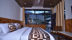 A bedroom with a bed, a chair, and a view of mountains through large windows. - Hotel Akash - Luxury Hotel In Mcleodganj (Dharamshala)