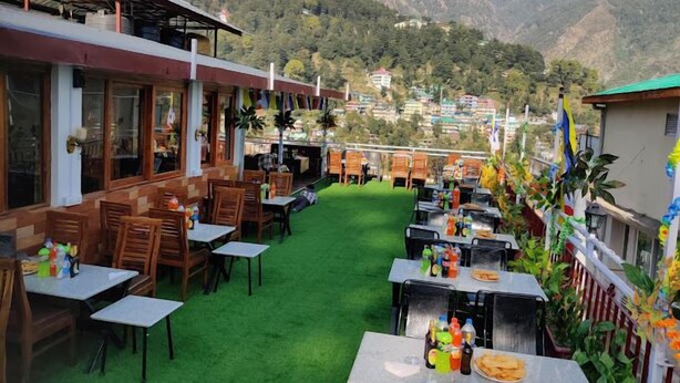 An outdoor restaurant with tables and chairs, a green artificial turf, and a mountainous backdrop. - Hotel Akash - Luxury Hotel In Mcleodganj (Dharamshala)