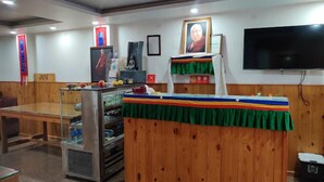A room with a wooden table, a refrigerator, a television, and a wall-mounted shelf with a framed picture. - Hotel Akash - Luxury Hotel In Mcleodganj (Dharamshala)