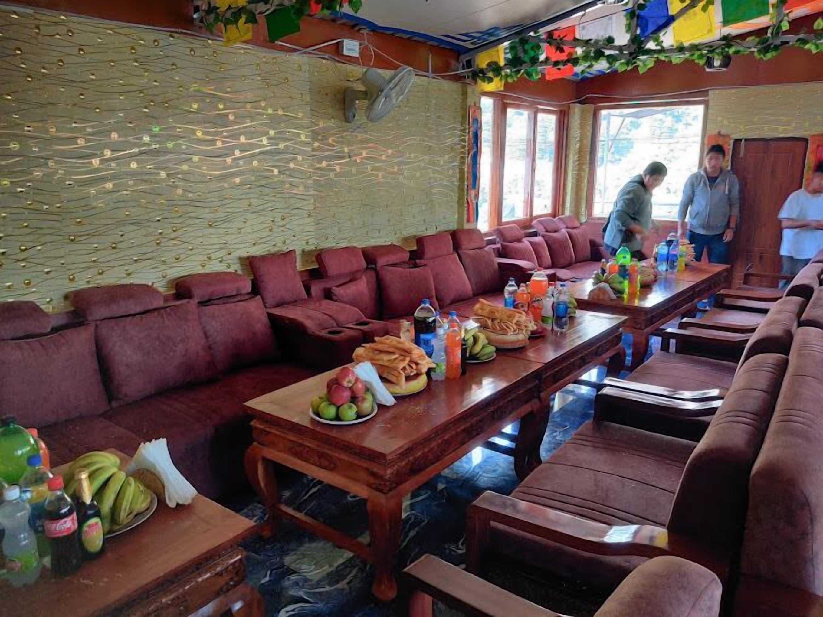 A dining area with a long wooden table set with fruits, snacks, and drinks. There are multiple cushioned seating areas and a wall with a textured pattern.