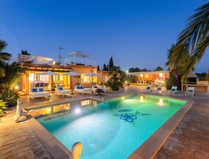 Un espace piscine avec des chaises longues et un coin repas, le tout entouré de palmiers et d’une maison moderne.