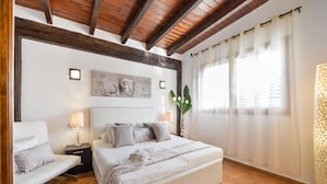 Een slaapkamer met een groot bed, een nachtkastje, een stoel, een kunstwerk aan de muur en een raam met gordijnen.