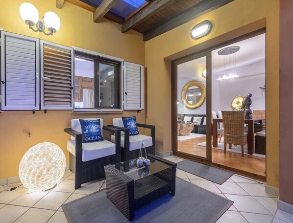 Une terrasse ensoleillée avec une porte vitrée donnant sur une salle à manger intérieure, des meubles en osier et une applique murale décorative.