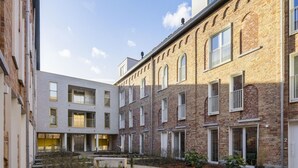 Une cour intérieure abritant un bâtiment moderne et un bâtiment traditionnel en briques, avec un jardin central agrémenté de sentiers et de petites plantes.