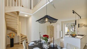 Un coin repas avec une table en bois foncé dressée pour six personnes, un lustre et une vue sur la cuisine et l’escalier.