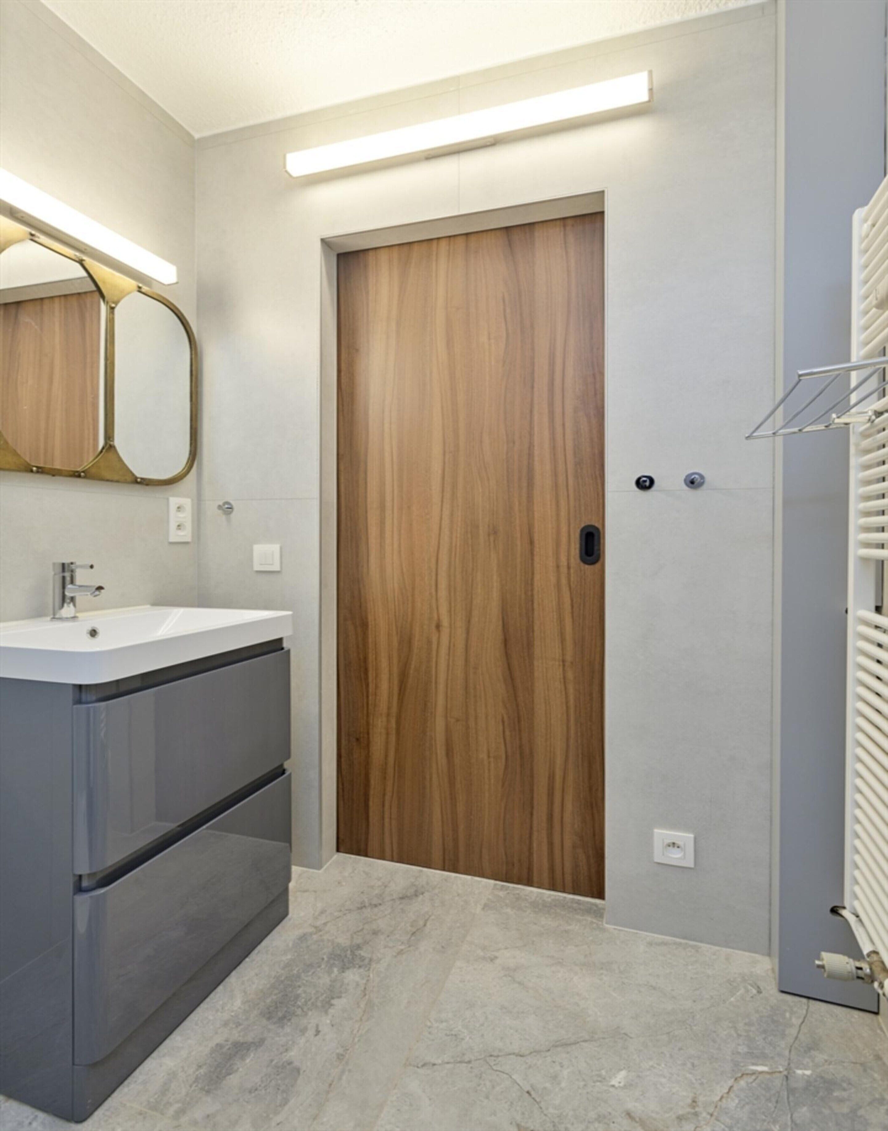 Un baño moderno con una puerta de madera, un lavabo blanco y un espejo con marco dorado.